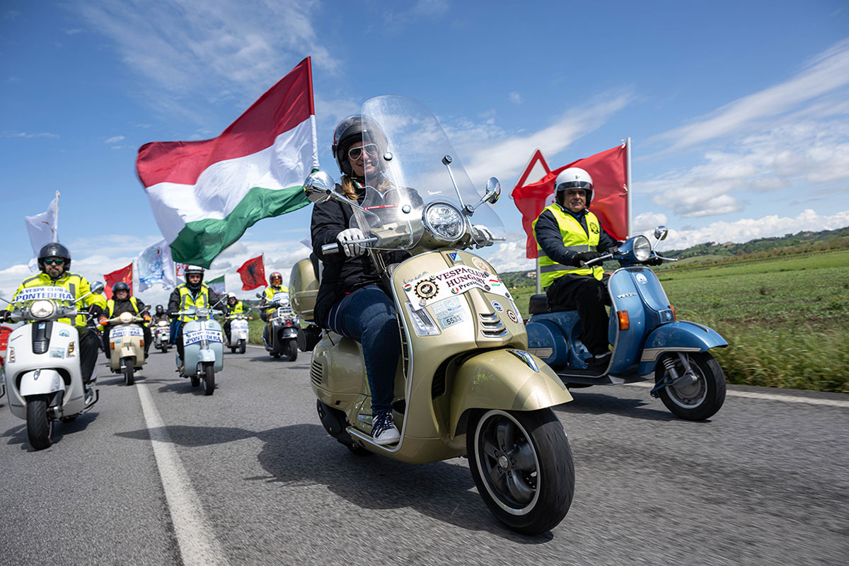 A parade of Vespa scooters with flags driving down a road during a previous celebration.